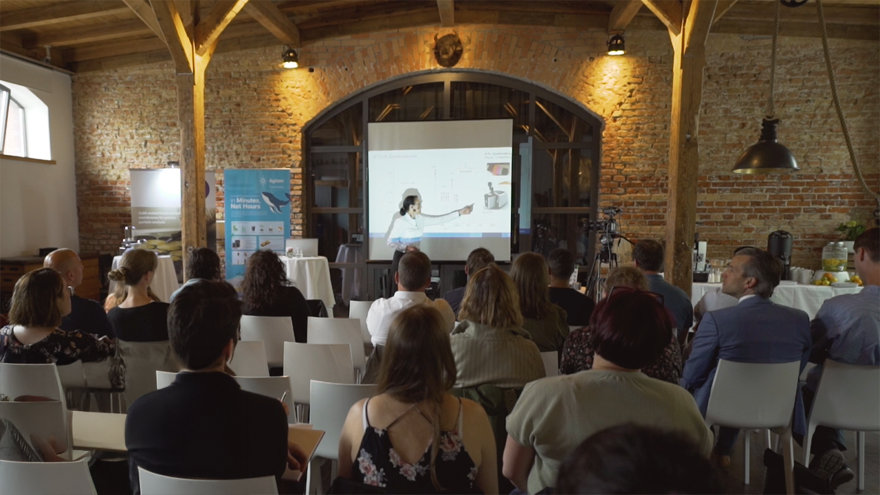 Photo of a speaker in front of a seated group pointing at an projected image